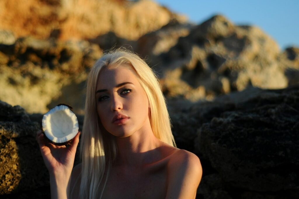 A blonde woman holds a coconut shell outdoors in warm sunlight, creating a natural and exotic atmosphere.
